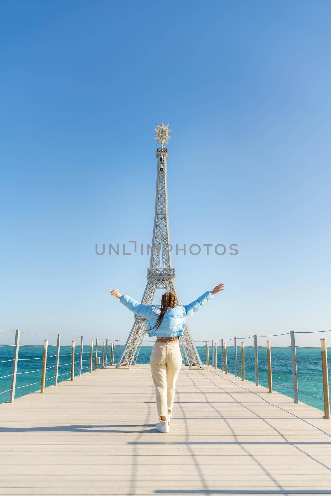 Large model of the Eiffel Tower on the beach. A woman walks along the pier towards the tower, wearing a blue jacket and white jeans