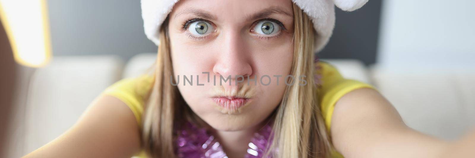 Portrait of blonde woman make funny facial expression, trying to scare, girl blow cheeks. Young female in hat and decor on neck, celebrating new year. Selfie, fun, party, christmas concept