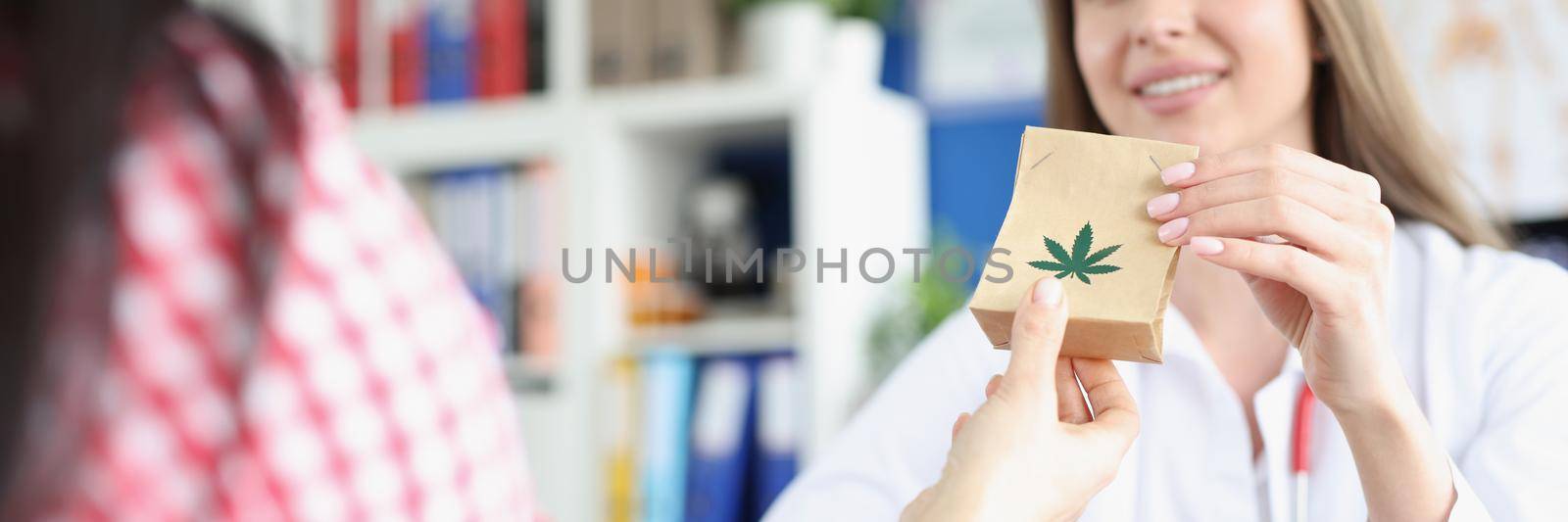 Portrait of smiling practitioner giving package with medical marijuana to female patient. Paper bag with cannabis sign. Narcotic pain relievers concept