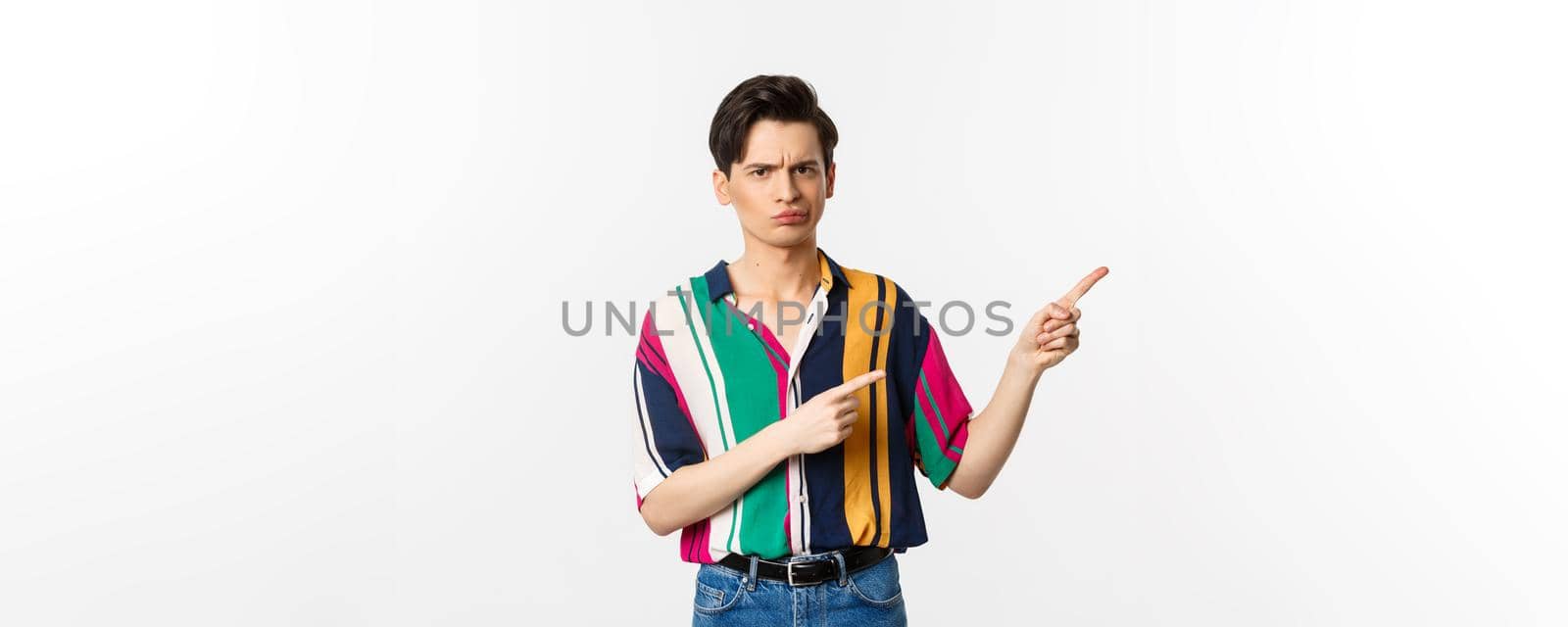Skeptical and displeased young man having concern, poitning fingers right and looking with disapproval, standing over white background by Benzoix