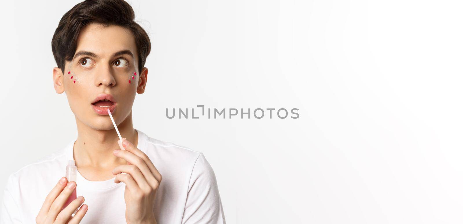 Close-up of beautiful androgynous man with glitter under eyes applying lip gloss, looking dreamy at upper left corner, white background.