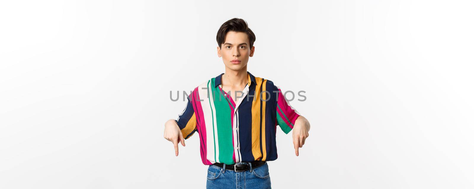 Young handsome queer guy pointing fingers down, showing advertisement and looking serious, standing over white background.