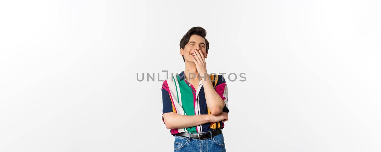 Image of handsome young man laughing over joke, having fun, cover mouth with hands and chuckle, standing over white background by Benzoix