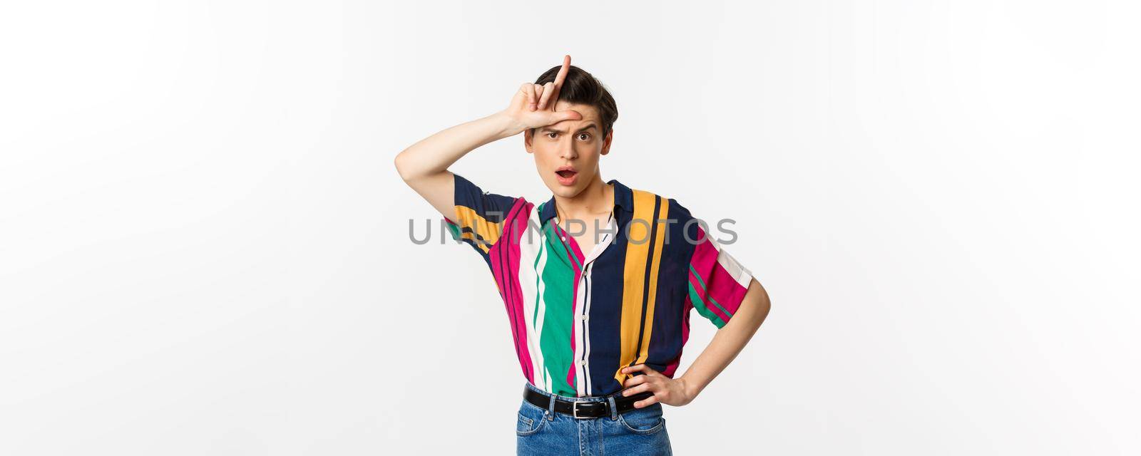 Sassy and arrogant guy showing loser sign on forehead, mocking someone, standing over white background.