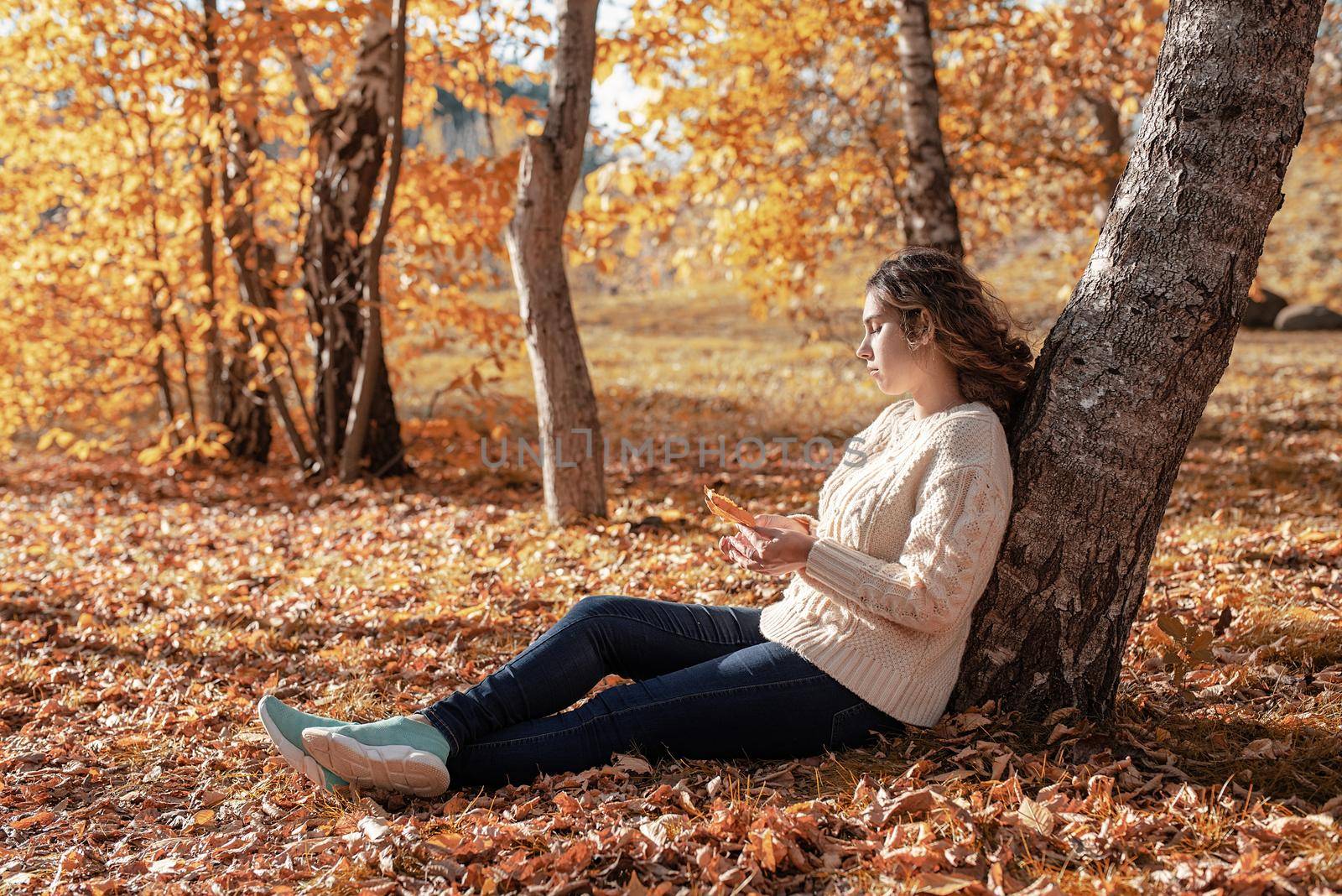 Young thoughtful woman sitting by the tree in autumn forest by Desperada