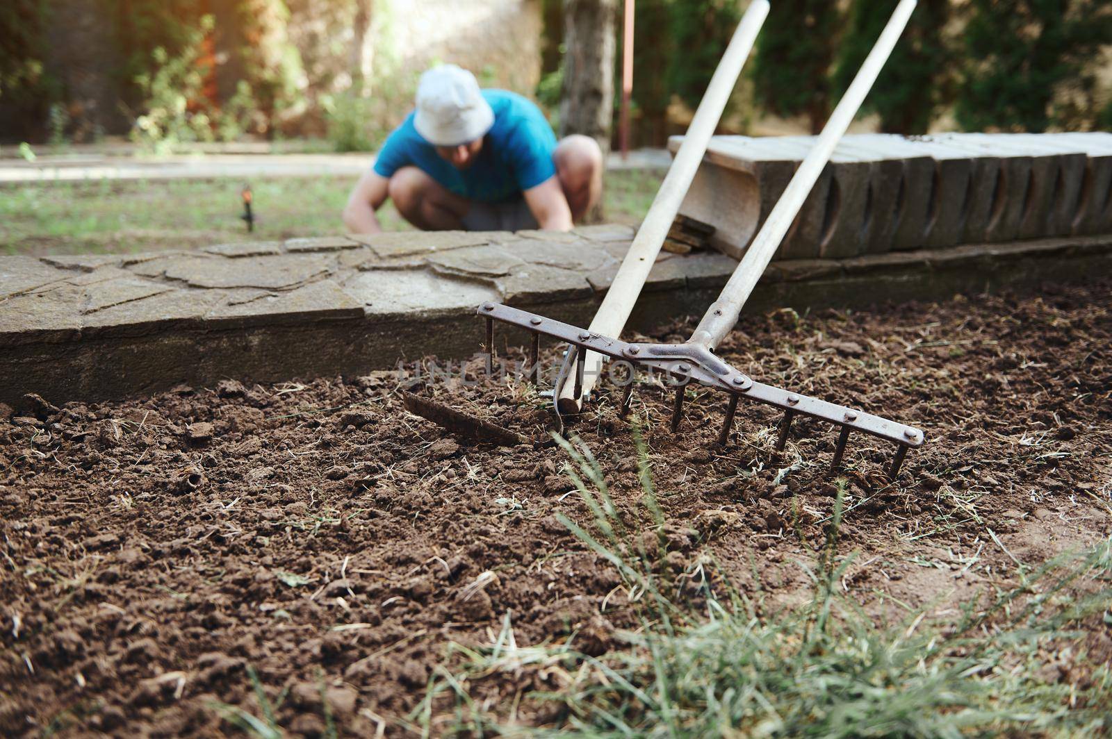 Focus on a garden shovel and rake on the loose ground and farmer working on the field on blurred background. Agribusiness. New agricultural planting season. Agro Industry. Agriculture, organic farming