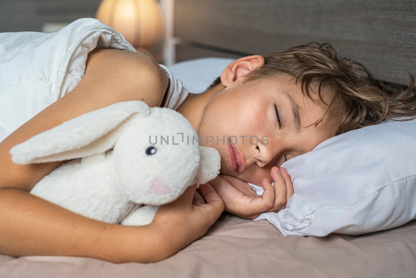 cute boy sleeping in bed with lamp. School child dreaming and holding plush toy.