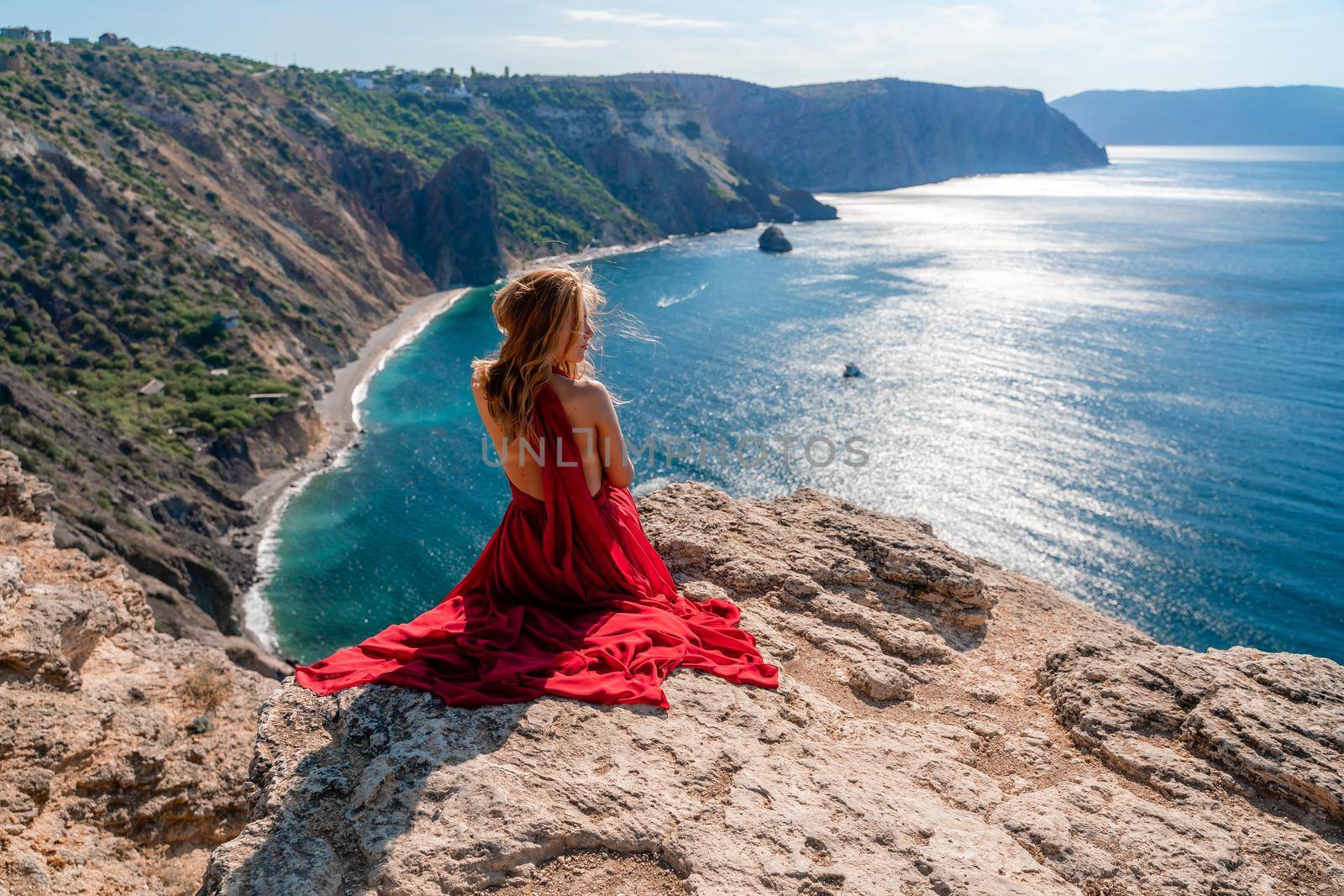 A girl with flowing hair in a long red dress sits on a rock above the sea. The stone can be seen in the sea