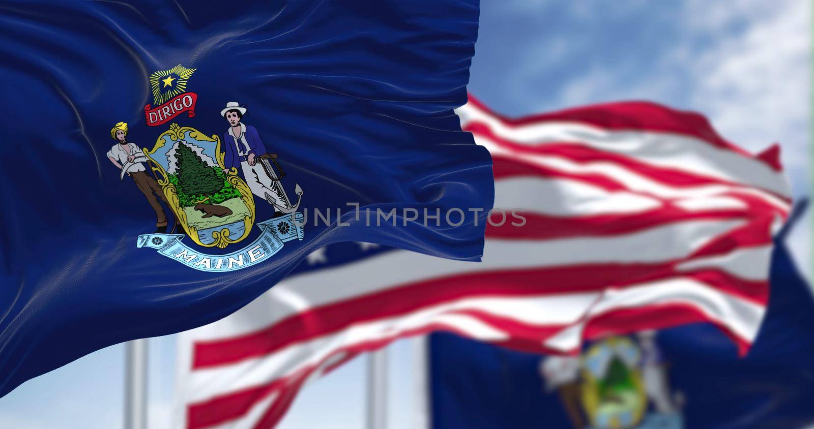 The Maine state flag waving along with the national flag of the United States of America. In the background there is a clear sky. Maine is a state in the New England region of the United States
