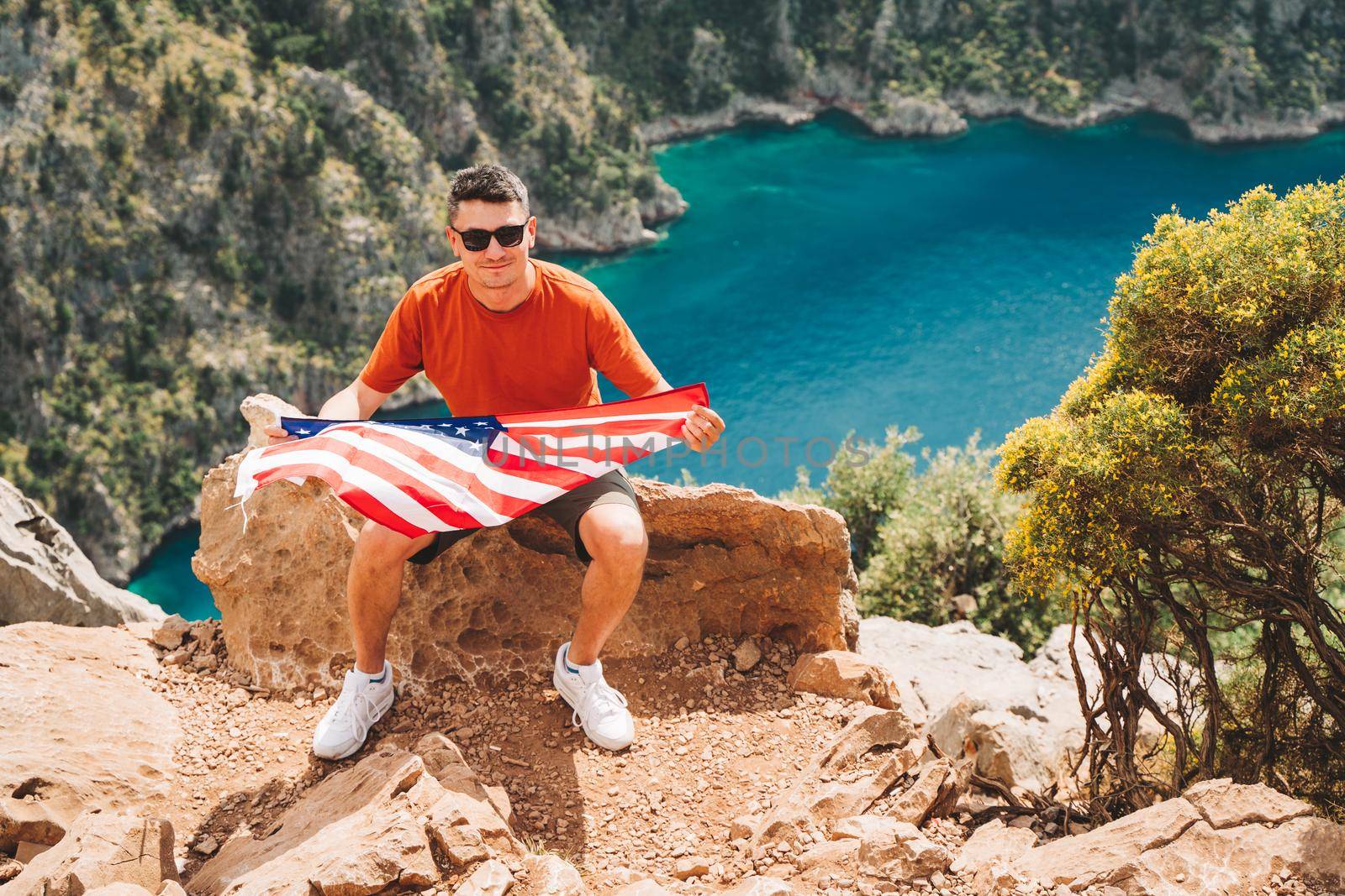 Young man sitting on a mountain top and holding the US flag in his hands. Veteran traveller holding the American flag while sitting on a mountain top. 4 July Independence Day.