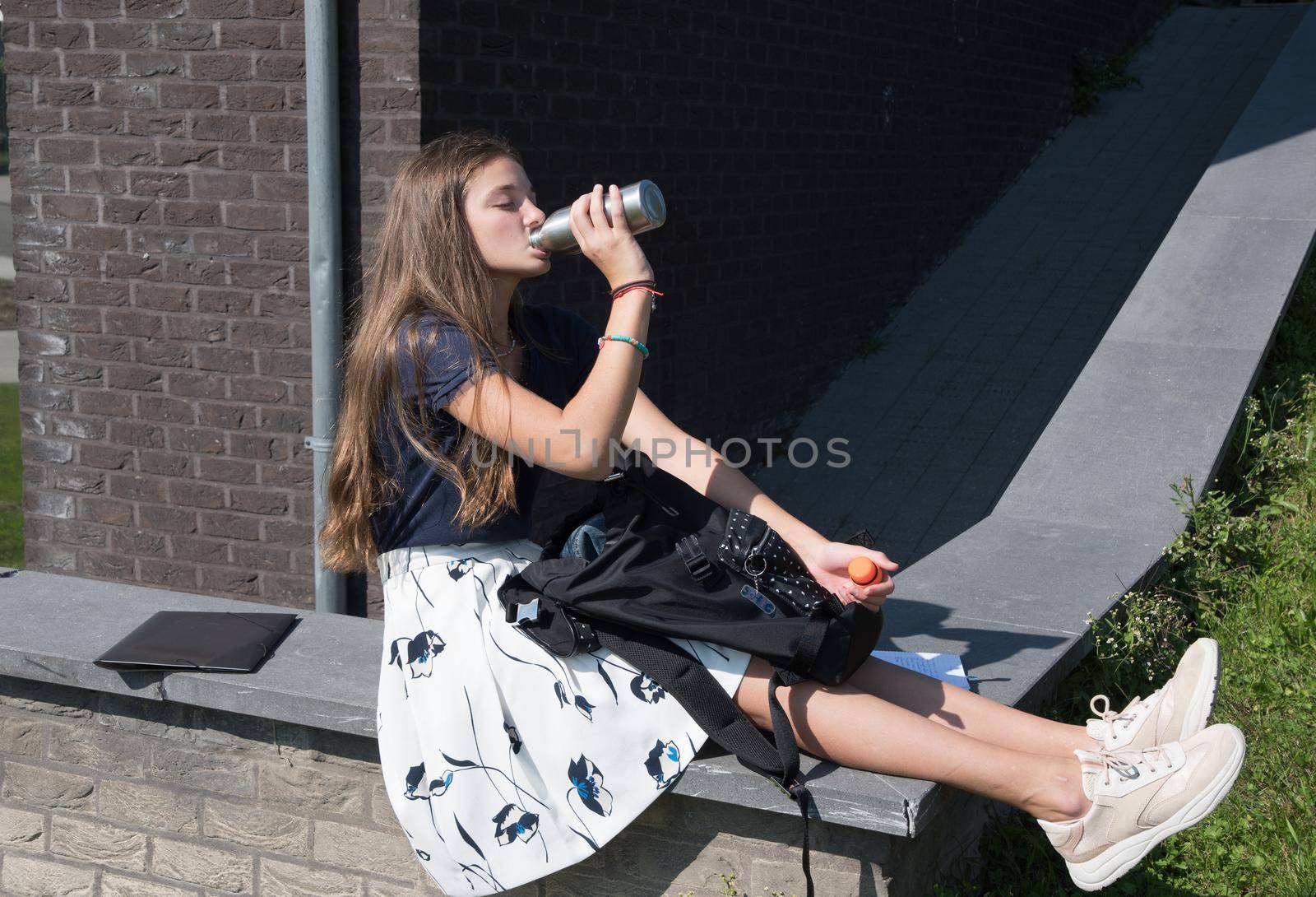 a teenage girl drinks water from a metal flask on a break between lessons and sits on a concrete curb near the school. High quality photo