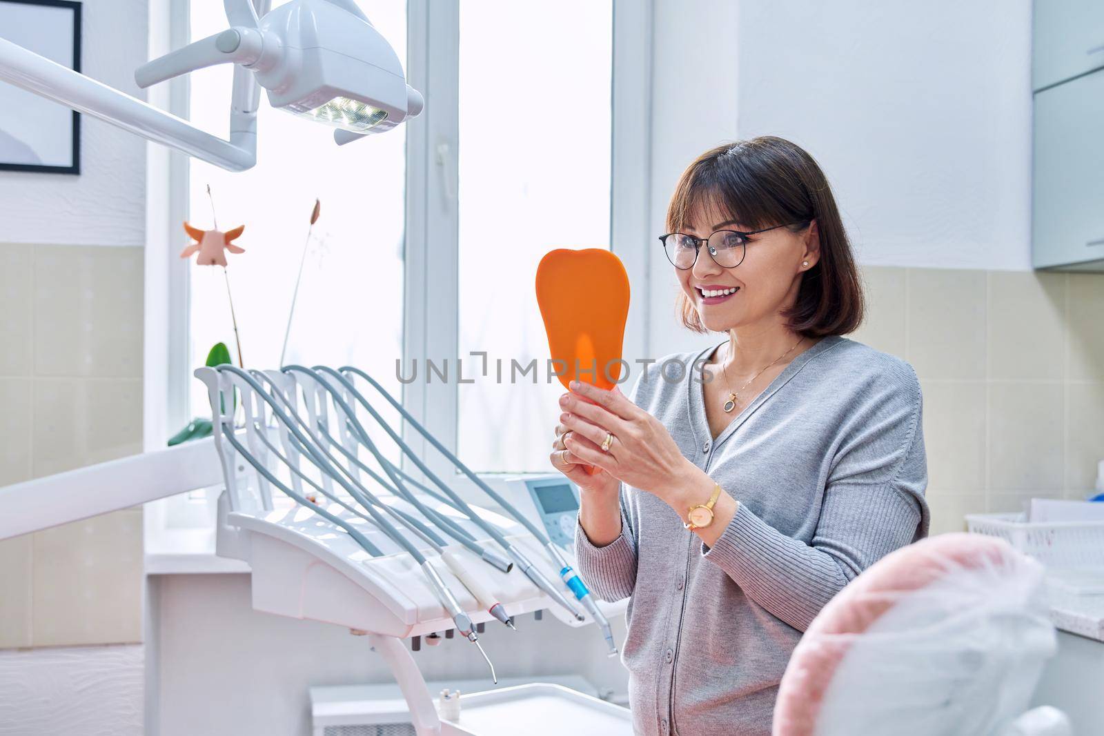 Happy middle aged woman dentist patient with a mirror in her hands looking at her teeth. Treatment, whitening, implants, medicine, health care and beauty smile concept