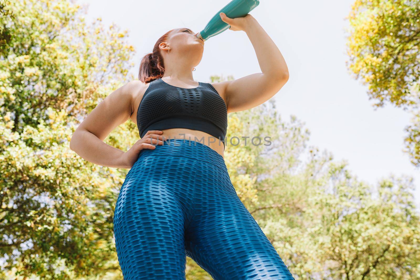 image from below of a young athlete drinking water to refresh herself from a bottle after a sports training. girl practising sport outdoors. health and wellness lifestyle. by CatPhotography