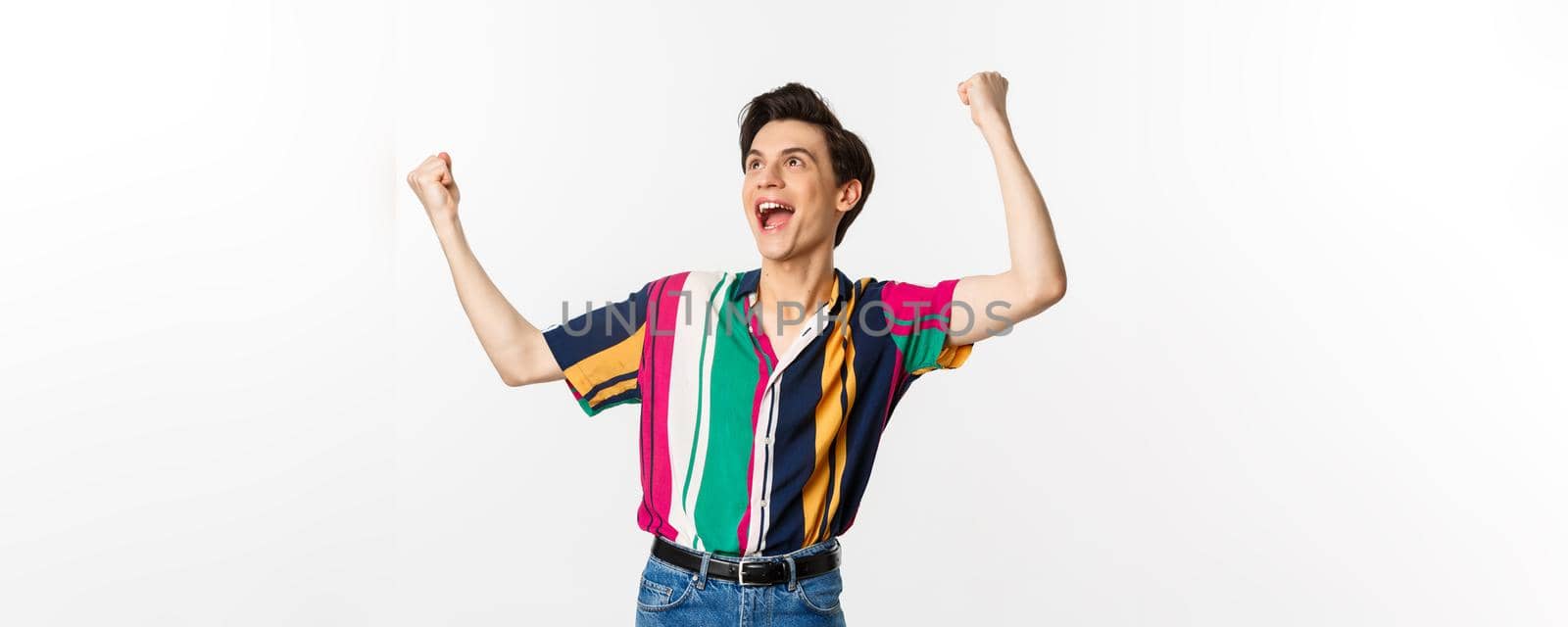 Happy gay man celebrating victory, raising hands up and rejoicing of winning, tirumphing while standing over white background by Benzoix
