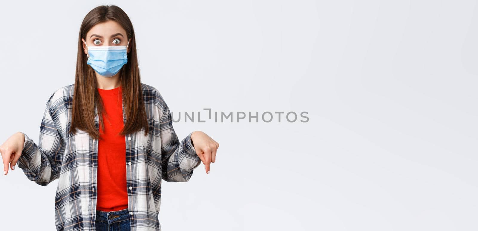 Coronavirus outbreak, leisure on quarantine, social distancing and emotions concept. Shocked and impressed young woman in medical mask and casual outfit, pointing fingers down, stare camera.