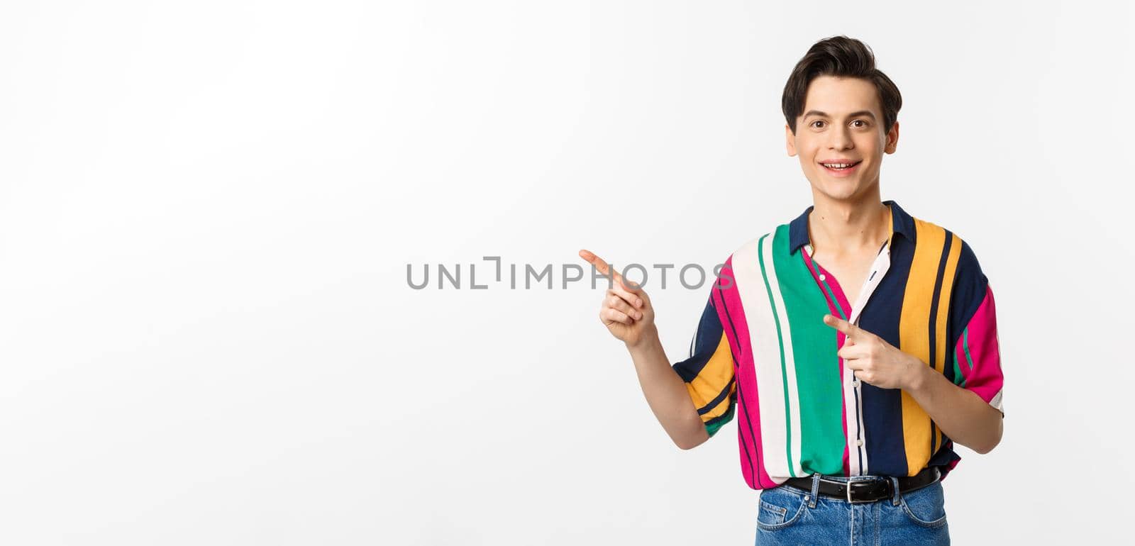 Portrait of handsome young man in stylish clothes, pointing fingers left and smiling, showing advertisement, standing over white background.