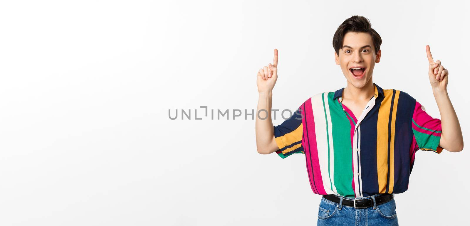 Happy attractive gay man pointing fingers up, smiling excited, showing top offer, standing over white background.