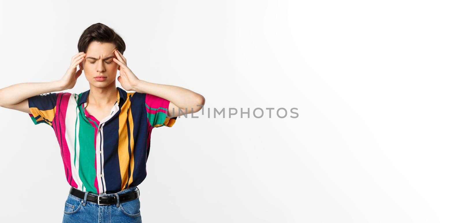 Image of young man having headache, looking troubled, holding hands on head, standing over white background.