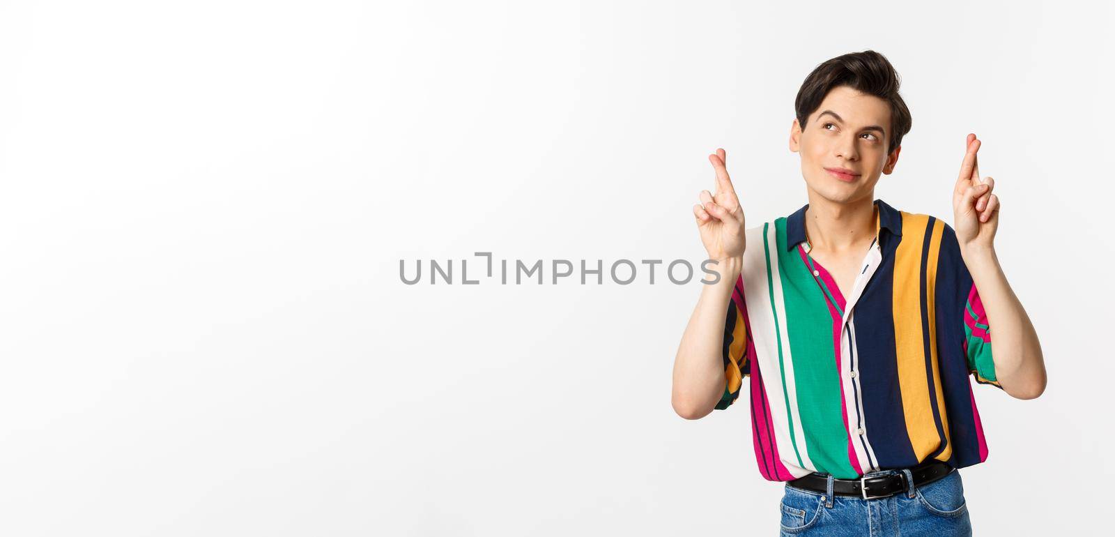 Dreamy young man making wish with fingers crossed, smiling and looking at upper left corner thoughtful, standing over white background by Benzoix