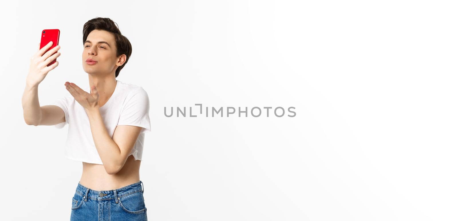 Beautiful gay man in crop top sending air kiss at phone camera, taking selfie or video chat on smartphone, standing over white background by Benzoix