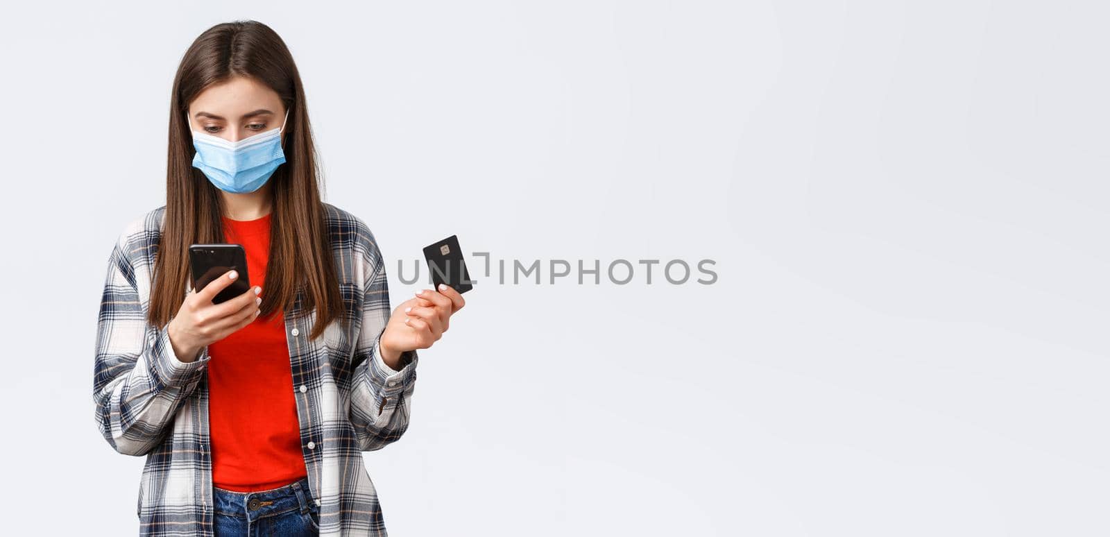 Coronavirus outbreak, working from home, online shopping and contactless payment concept. Girl in medical mask holding credit card, order clothes using mobile phone application.