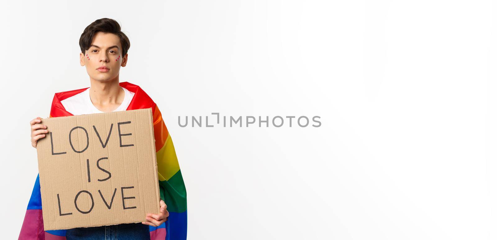 Serious and confident gay man wearing rainbow lgbt flag, holding sign for pride parade, standing over white background.