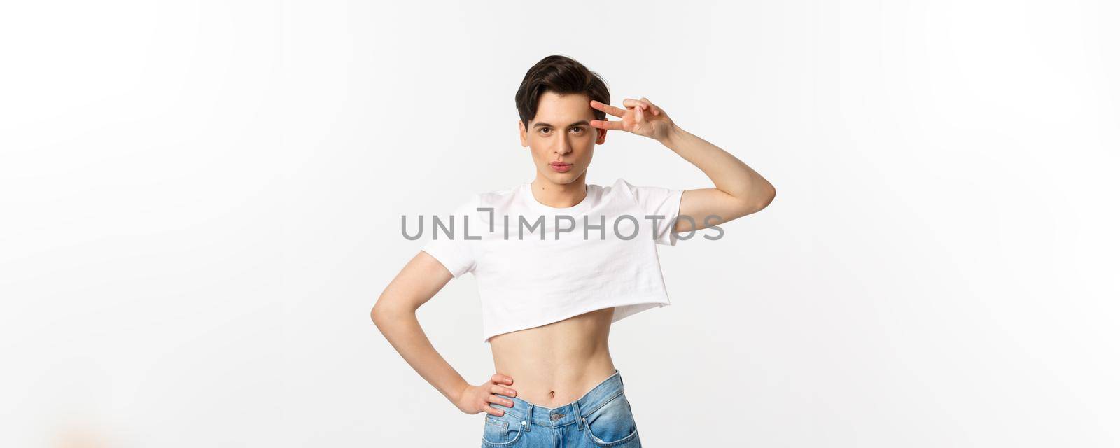 Beautiful androgynous man in crop top showing peace sign and smiling, standing over white background.