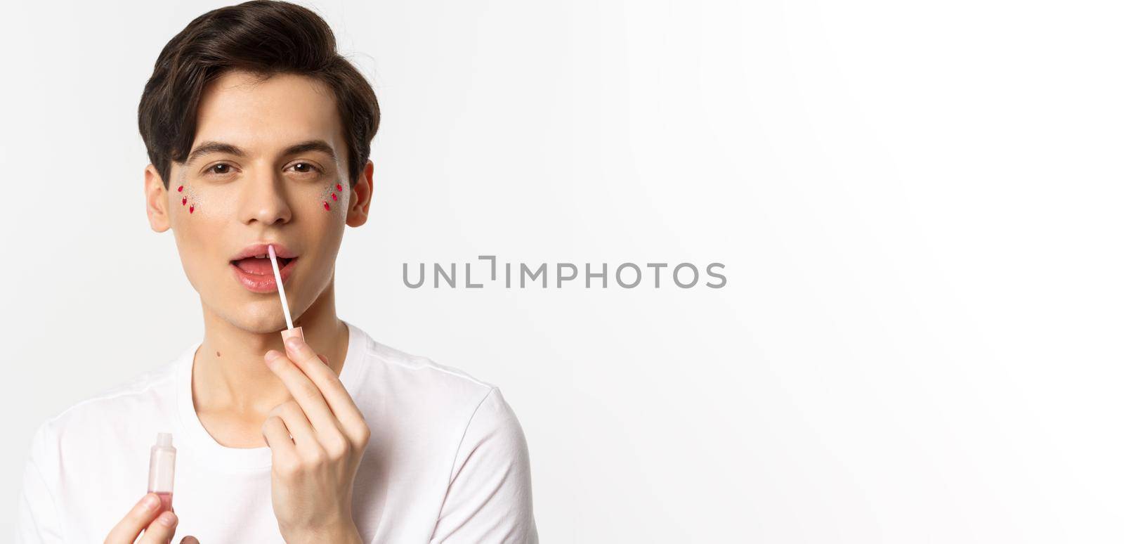 Close-up of gender fluid person with glitter on face, applying lip gloss lipstick and looking at camera, standing against white background by Benzoix