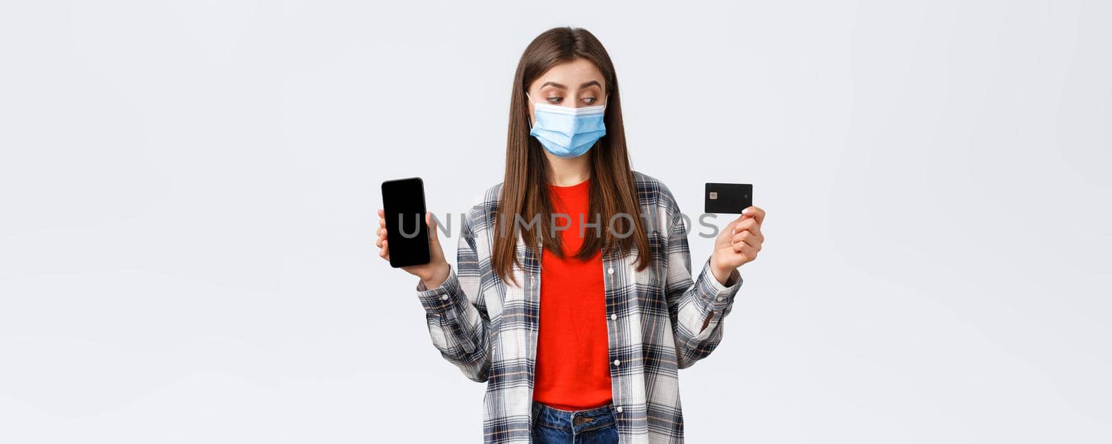 Coronavirus outbreak, working from home, online shopping and contactless payment concept. Girl in medical mask look at credit card as show mobile phone screen, making order.