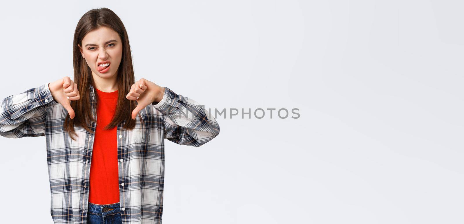 Lifestyle, different emotions, leisure activities concept. Displeased and unimpressed young picky girl in checked shirt, thumb-down and show tongue to express dislike, rate awful movie by Benzoix