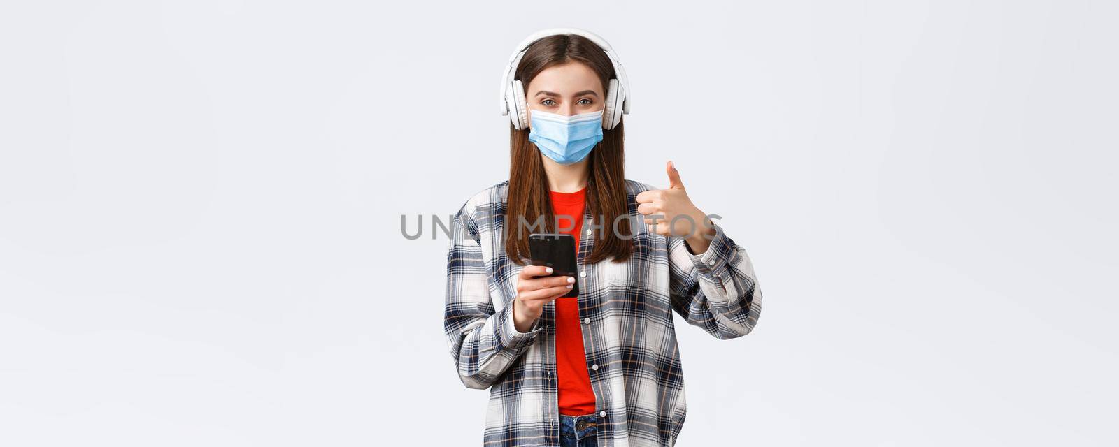 Social distancing, lifestyle on covid-19 outbreak, coronavirus concept. Cheerful pleased good-looking woman in medical mask, approve nice sound of wireless headphones, thumb-up, hold phone by Benzoix