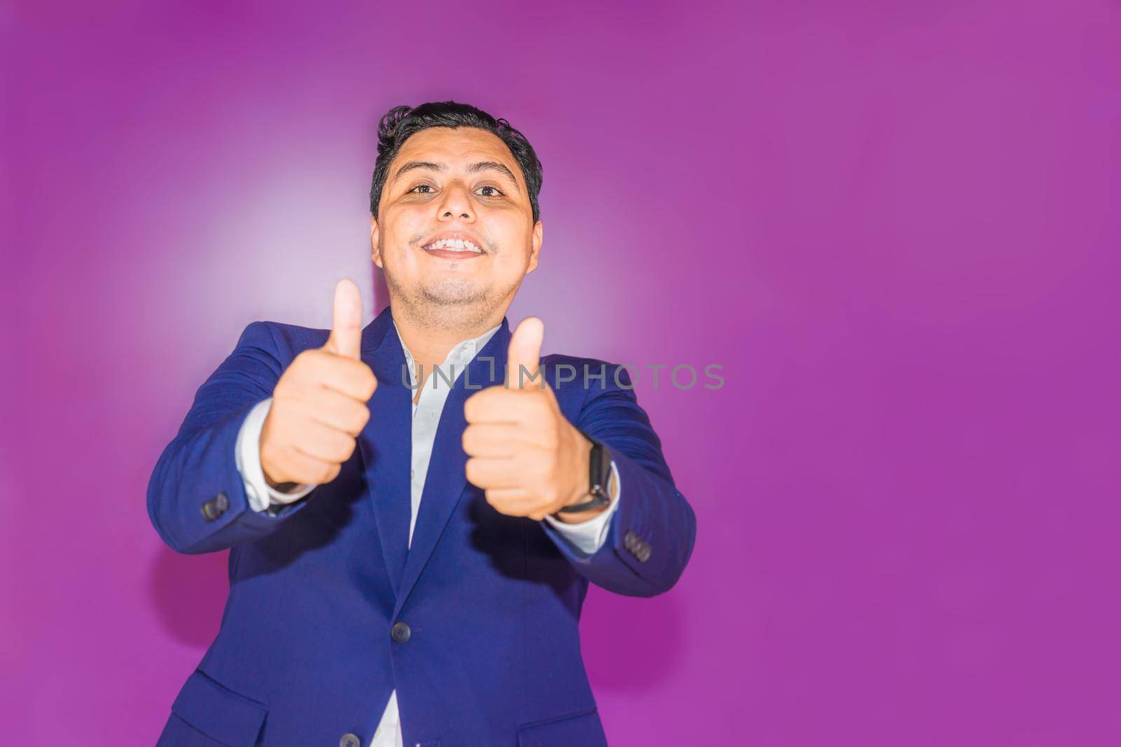 Latin man in a blue suit raising the thumbs of both hands on a plain purple background by cfalvarez
