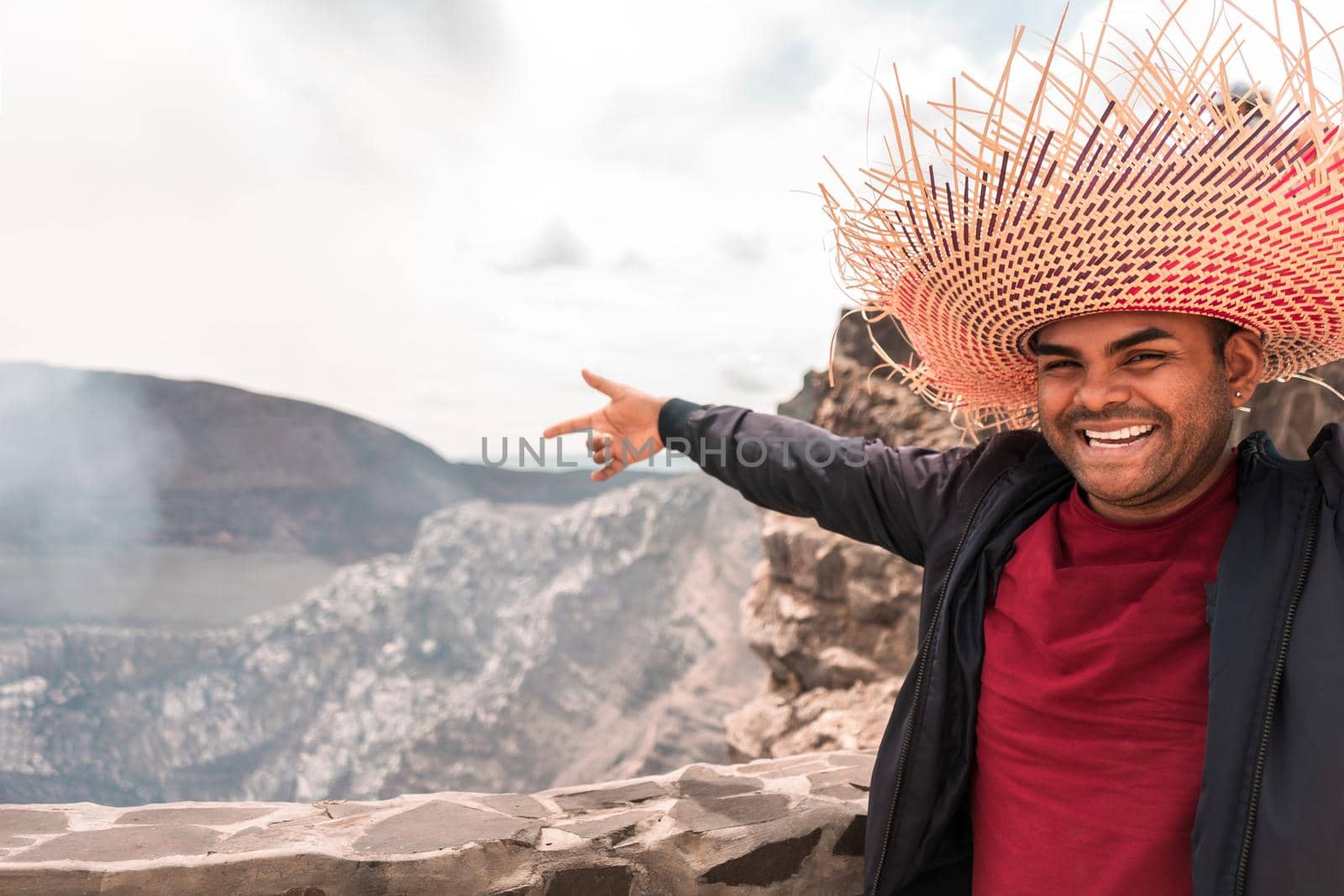 Portrait of a mestizo latin man looking at camera and pointing to the crater of a volcano wearing a colorful travel hat in an exotic tourist destination in Masaya, Nicaragua. Photo with copy space.