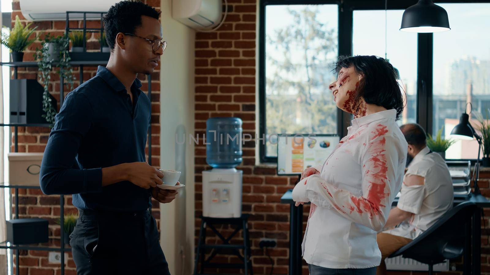 Hungry creepy zombie talking with businessman while dead coworker covered in blood walks by. Person discussing with bizarre looking evil undead monster in office while mindless colleague passes by.
