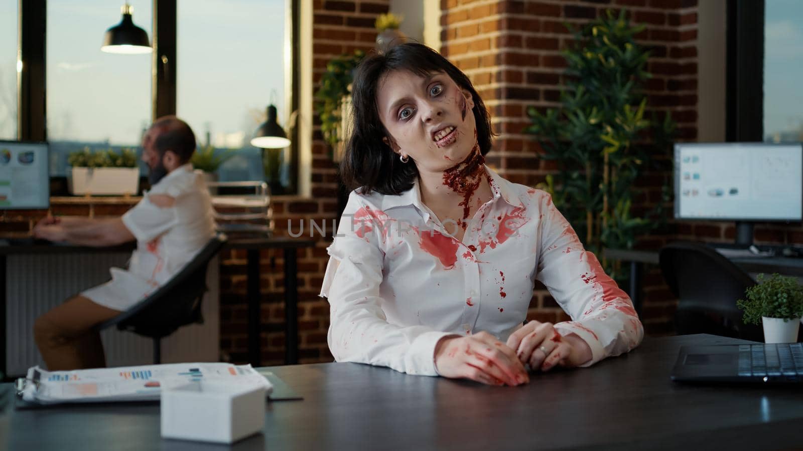 Scary zombie woman with bloody deep wounds in office waving hand at camera. Brain-eating creepy looking monster sitting at table in workspace while looking brain dead and evil.