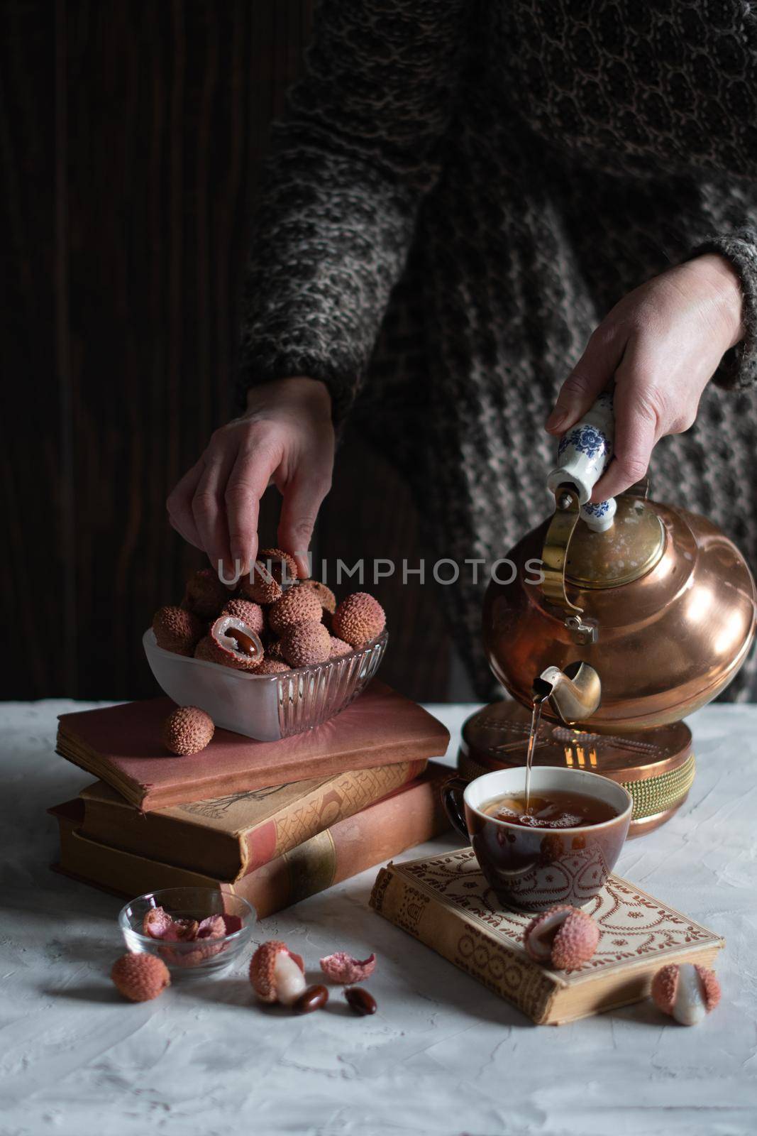 A woman pours tea from a copper teapot into a cup against the background of a stack of books and a cup with lychee, homely cozy tea drinking and reading,. High quality photo