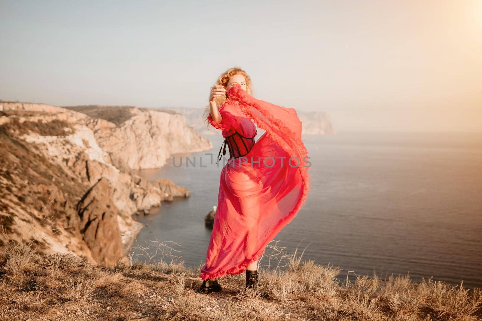 Side view a Young beautiful sensual woman in a red long dress posing on a volcanic rock high above the sea during sunset. Girl on the nature on blue sky background. Fashion photo