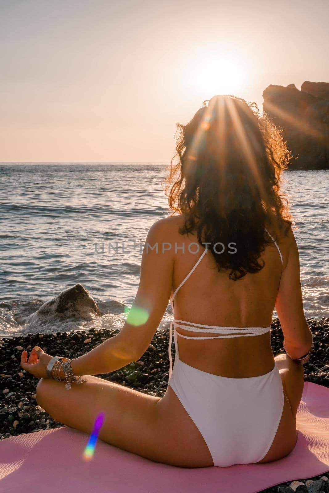 Young woman in swimsuit with long hair practicing stretching outdoors on yoga mat by the sea on a sunny day. Women's yoga fitness pilates routine. Healthy lifestyle, harmony and meditation concept.