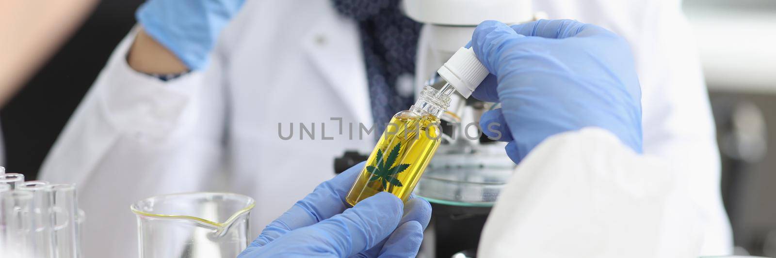 Close-up of persons hand hold jar with cannabis oil and going to add drop under microscope. Woman scientist explore sample, protective uniform. Lab concept