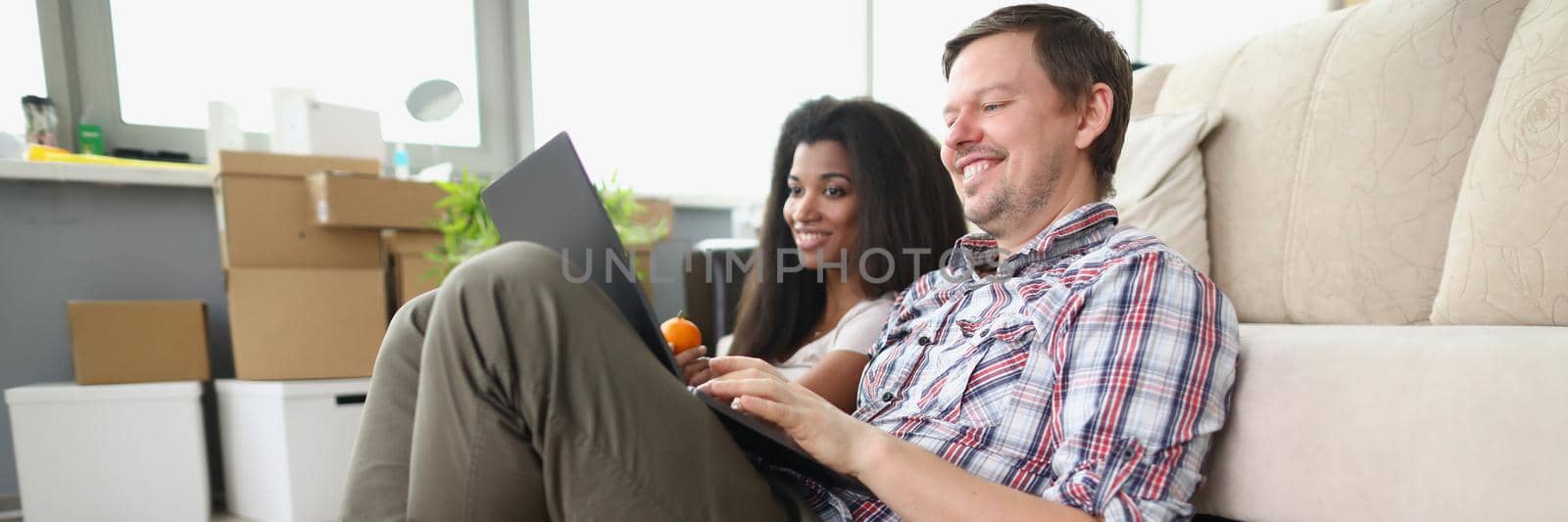 Low angle of cheerful smiling couple watch show online on laptop at home. Man and latin girl sit on floor, spend time on weekend. Relationship, fun concept