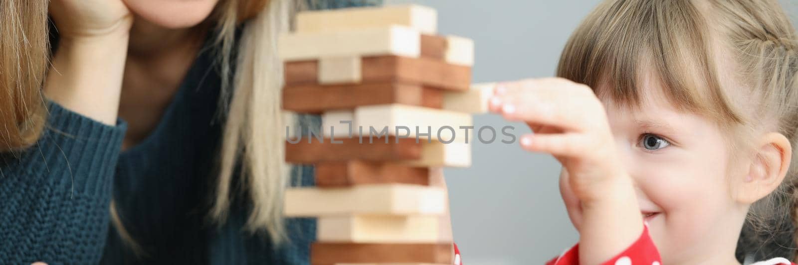 Portrait of beautiful little girl kid pull wooden block out of high tower and smile. Mum join daughter in playing board game. Parenthood, childhood concept