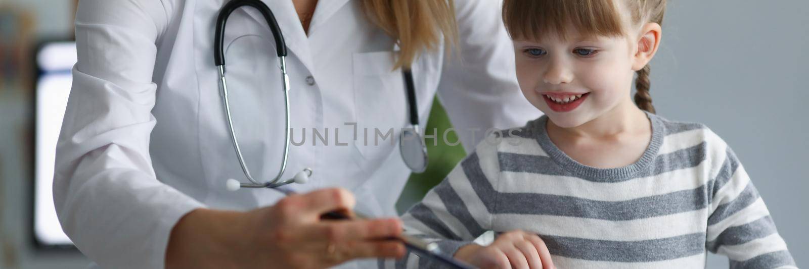 Portrait of pediatrician female try to distract child attention and make diagnostic. Smiling kid on planned checkup in clinic. Medicine, healthcare concept