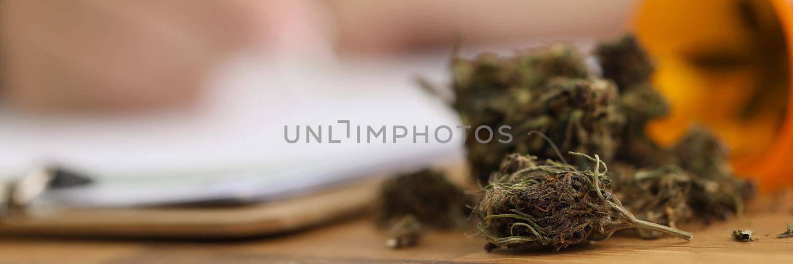 Close-up of dried pieces of marijuana on doctors desk fell out of plastic bottle. Doctor on background write prescription for patient. Hemp, health concept