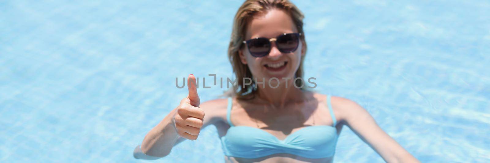 Portrait of happy young woman swim in pool and show thumbs up from water. Relaxing atmosphere on vacation, sunny day. Holiday, resort, hotel, fun concept