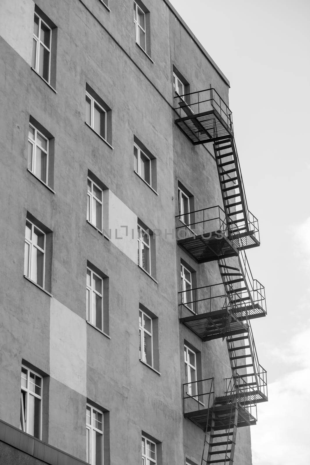 Silhouette of a fire escape on a high-rise building against a blue sky with clouds. Some of the stairs are broken. There is free space for text. Black and white photo