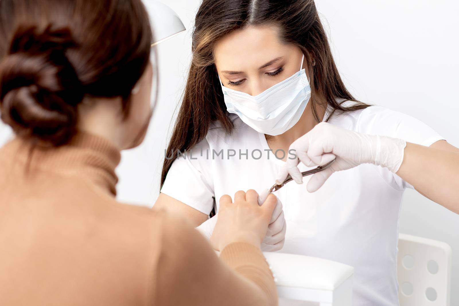 Manicurist in protective mask using manicure cutter to remove cuticle of female nails in manicure salon