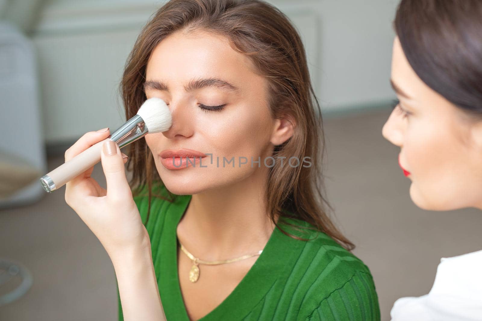 Makeup artist applying dry cosmetic tonal foundation on nose of young caucasian woman using brush tool in beauty salon