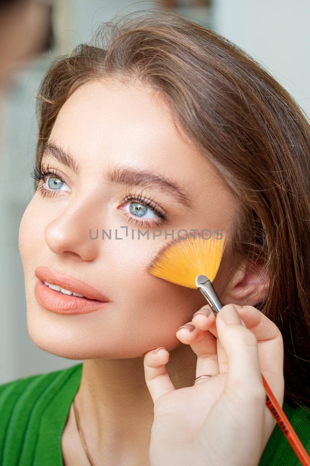 Professional make up artist applying powder by brush on cheeks of beautiful young caucasian woman in beauty salon