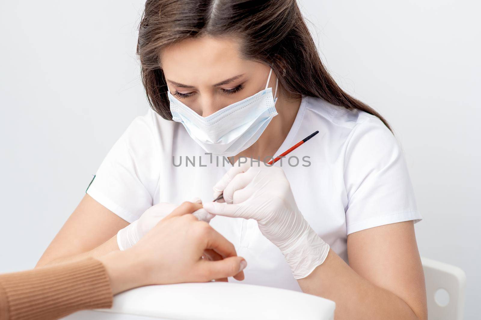 Manicure master with protective mask applying transparent nail polish on female nails in beauty salon