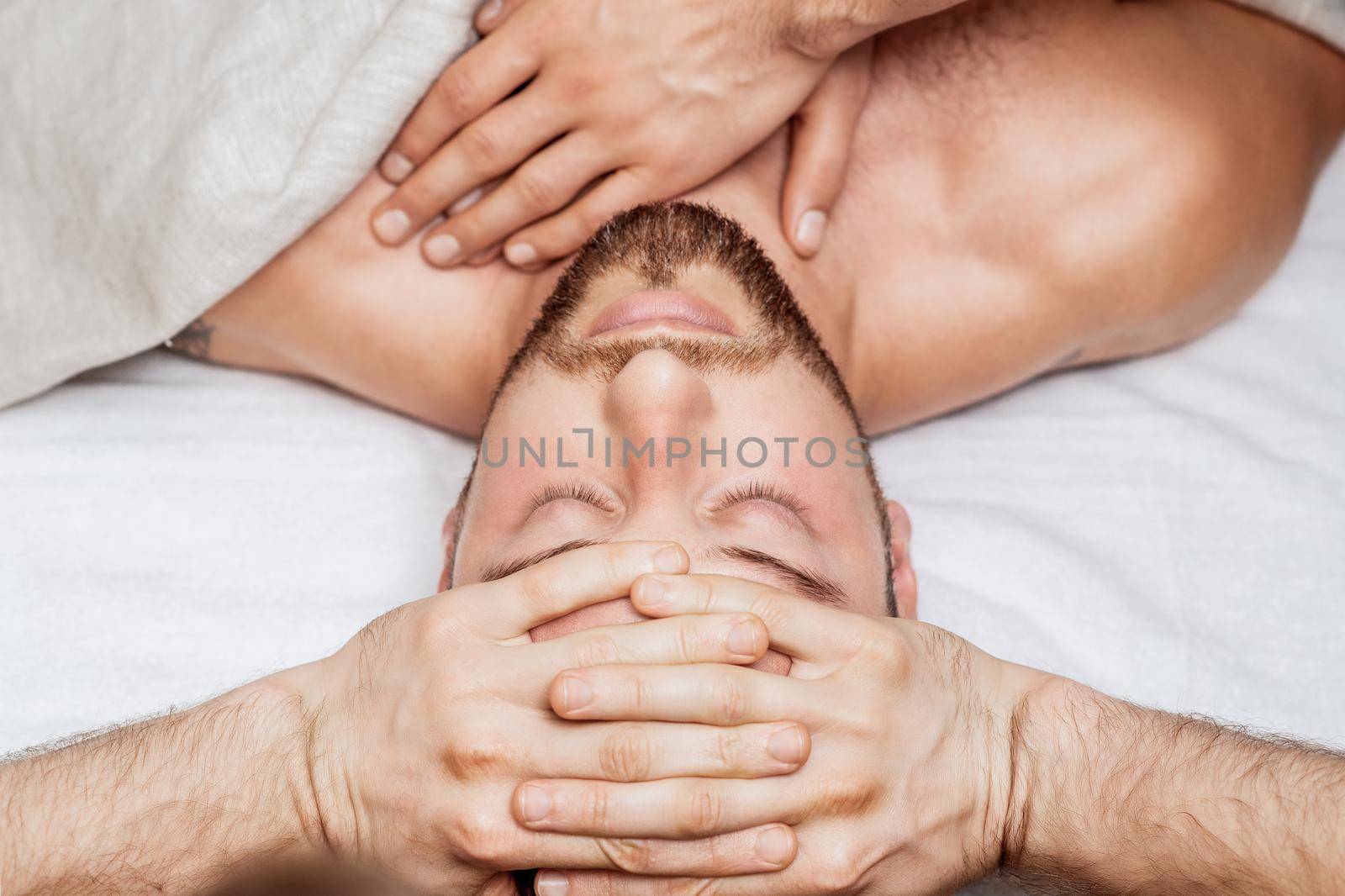Handsome young man receiving relaxing head massage by four hand of two masseurs in health spa center
